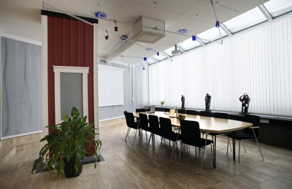 Bild på konferenslokalen Oasen. På bilden syns ett avlångt konferensbord med svarta stolar. Rummet har ett fönster längs ena långsidan. Fönstren är skymda med gardiner. Rummet har även en bred pelare av trä, designad som en liten stuga med röd panel och vita knutar.