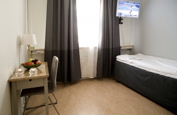 Bild på ett hotellrum. På bilden syns en enkelsäng, en tv på väggen och ett skrivbord med tillhörande stol.