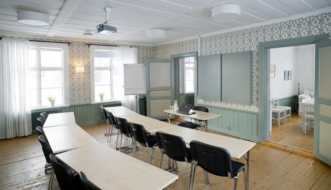 Konferensrum i byggnaden Gammelgård. Två rader med avlånga bord och svarta stolar. Alla stolar är riktade mot talarens plats. Rummet har gamla trägolv, turkos gammal tapet på väggarna och vitt tak. Rummet är litet men känslan i bilden är mysig.